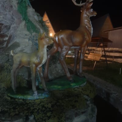 This image depicts a nighttime scene with two statue figures of deer, one larger and one smaller, positioned in what appears to be an outdoor artificial landscape. The larger deer statue, with antlers, is brown and appears to be an adult. The smaller deer statue, a fawn, is also brown and positioned to the left of the adult. They are standing on a green surface, possibly representing grass, which is part of a diorama with a rock formation on the left. In the background, there are wooden structures and a fence, suggesting an outdoor park or entertainment area. The lighting suggests it is dark, with artificial light illuminating the statues. The location is identified as Sosnovka, Kyrgyzstan. There are no people visible in the image.