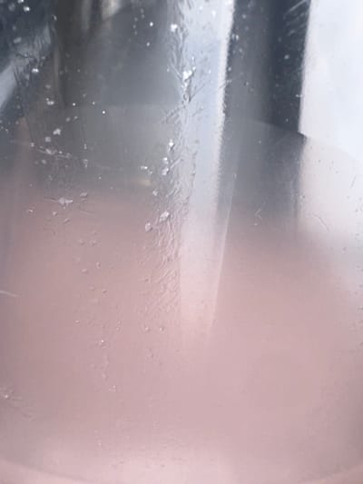 The image shows a close-up view of a surface that appears to be made of glass, possibly a window or windshield. The surface is covered in a thin layer of frost or ice, with intricate crystalline patterns formed by the freezing process. There are also small water droplets or ice crystals scattered across the surface. The background is out of focus, but suggests an outdoor scene with soft, diffused light, possibly indicating overcast weather. The overall impression is one of cold and stillness. The image does not contain any people, text, or obvious location cues other than the provided context of Letterkenny, Ireland. The time of day is indeterminate, but the frost suggests a cold, likely early morning or late evening during a colder season.