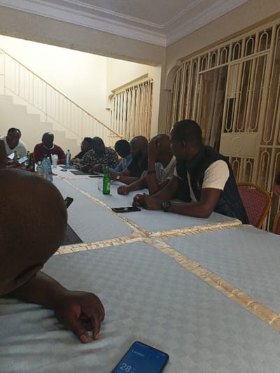 A group of eight men are seated around a long table covered with a light blue tablecloth with a gold trim. The table is cluttered with drinks and cell phones. A man in a dark vest is looking toward the end of the table. In the background, there is a staircase and a barred window. A cell phone on the table in the foreground shows the time and temperature in London.