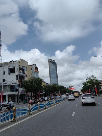 The image captures a daytime street scene in Thuận Hóa Ward, Vietnam, under a sky with prominent white and grey cumulus clouds and patches of blue. The viewpoint is from a low angle on an asphalt road featuring white lane markings. A blue and white patterned metal barrier, showing some rust, delineates the left side of the road, separating the main vehicular lanes from an area with parked motorcycles and a sidewalk.
Along the left side, a row of multi-story buildings with varied architectural styles line the street. These include commercial establishments on the ground floor, indicated by Vietnamese signage such as "NHÀ XUẤT DƯ" and "ANH KHÁNH". Green-leafed trees are planted along this stretch. A tall, slender, glass-clad skyscraper with a distinct, possibly trapezoidal, shape dominates the mid-background, featuring a logo near its apex. To the far left, a metal lattice tower is partially visible.
Traffic on the road moves away from the viewer. A white SUV with a visible license plate segment '75A-XXX.XX' occupies the right lane in the foreground. A silver sedan is positioned in the left lane ahead of the SUV. Further down the street, a light-colored bus and additional vehicles are visible. Numerous motorcycles are present along the left side of the road, behind the barrier, with several individuals wearing helmets. One motorcyclist in a white helmet and light-colored jacket is depicted in the immediate foreground, facing left. Red and white banners are affixed to poles along the street.