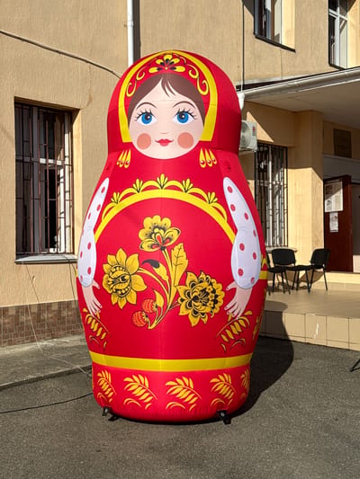 An oversized, inflatable matryoshka doll, predominantly red with yellow and white accents, stands inflated on an asphalt surface in front of a light brown, two-story building. The building features barred windows on the ground floor and regular windows on the upper floor, as well as an air conditioning unit and an entrance with a sign indicating "INFORMATION." Two black chairs are placed on a tiled step leading up to the entrance. The matryoshka doll is adorned with a painted face featuring blue eyes, rosy cheeks, and red lips, as well as traditional floral patterns. The scene appears to be outdoors, likely in a public space within the city of Progress, Russia.