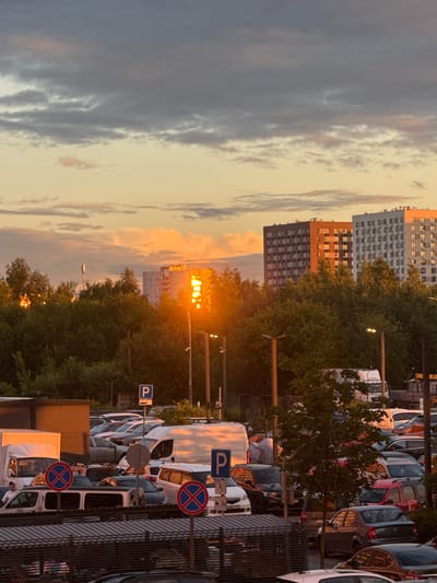 This is a vertically oriented photograph of a street scene in Drozhzhino, Russia, at dusk. The sun is low in the sky, casting a warm, golden light that illuminates the clouds and the windows of a multi-story building. The sky is filled with a mix of gray and pale yellow clouds, suggesting a transition from day to night.
In the foreground, a row of parked cars and vans line the street. Several of these vehicles are white vans, and others are passenger cars in various colors. Visible parking signs with the letter "P" are posted, indicating parking areas. Two circular red and blue traffic signs, likely indicating a prohibition, are also present. The vehicles are parked alongside a metal fence and some trees.
Behind the parked vehicles, dense green trees form a line along the street. Beyond the trees, several tall residential buildings are visible. One building is a striking orange color, contrasting with the gray and white facades of the buildings next to it. The windows of these buildings reflect the warm light of the setting sun.
Streetlights are also visible, some illuminated, contributing to the overall ambiance of dusk. The overall mood of the image is peaceful and serene, capturing the quiet beauty of a suburban street at the end of the day. No people are clearly visible in the frame.