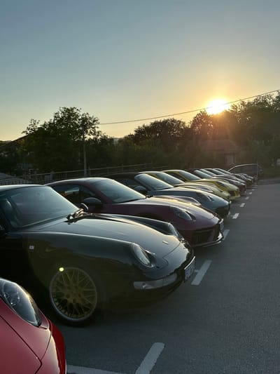 A line of six Porsche cars are parked in a lot at sunset. The cars are all different colors, including black, red, purple, and yellow. The sun is setting behind the trees in the background, casting a warm glow over the scene. The cars are parked in a row, with white lines marking the parking spaces. In the foreground, the front of a red car is visible. The cars appear to be in good condition. The location is Kerenite, Bulgaria.