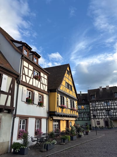 The image captures a street view in Colmar, France, likely in the historic Petite Venice district, characterized by its traditional Alsatian architecture. Several half-timbered buildings, painted in white and dark wood, line a cobblestone street. One building is prominently painted in bright yellow with dark timbering, featuring numerous windows with white shutters and flower boxes filled with pink and red blossoms. To the right, another half-timbered building in green and white is visible, and further down, a building with black timbering on a cream background. Planters with green foliage and white flowers are placed along the sidewalk, and outdoor seating with tables and chairs is set up in front of one of the establishments, suggesting a cafe or restaurant. Two individuals are seen standing near a sign that reads "LE QUAI 21". The sky above is a bright blue with scattered white clouds, indicating pleasant weather.