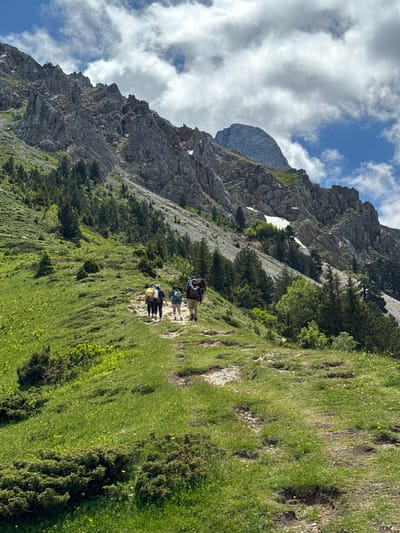 A group of four hikers with backpacks are walking on a dirt path that winds up a grassy mountainside. The path is flanked by lush green grass and scattered yellow wildflowers. In the background, a rugged, rocky mountain range rises steeply, with patches of snow visible on its slopes. The sky above is a mix of blue and white clouds. The scene appears to be in the natural landscape surrounding Jošanica, Montenegro.