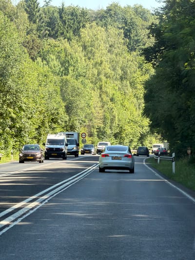 The image depicts a two-lane road winding through a densely wooded area, likely in Niederanven, Luxembourg. The road surface is asphalt, marked with double white lines. Several vehicles are traveling in the same direction, including a light-colored Tesla in the foreground, followed by a dark car, and then two vans and another car. The surrounding environment is characterized by lush green trees and foliage on both sides of the road, with a clear blue sky visible above. The scene appears to be captured during daylight hours, with sunlight casting shadows on the road. There are no visible pedestrians or any other notable human presence besides the occupants of the vehicles. The overall impression is one of a typical rural or suburban road scene in a heavily forested region.