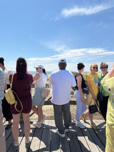 In Morskoye, Russia, a group of individuals stands on a wooden boardwalk, observing the scenic landscape under a clear blue sky with scattered clouds. The sun casts shadows on the weathered wood.
On the left, a person with dark hair featuring pink streaks wears a maroon dress and a yellow backpack. Beside them, a woman in a white tank top and light blue shorts looks to her right, a beige crossbody bag slung over her shoulder. She wears a white baseball cap and sunglasses.
To the right of the woman in blue shorts, a man in a white t-shirt and grey pants faces away from the camera. Further right, a woman in a black top and striped pants is visible. Next to her, a woman in a yellow t-shirt and pants, wearing sunglasses, holds a dark handbag.
Another woman, to the far right of the yellow-shirted woman, wears a green patterned top and sunglasses, also holding a dark handbag. In the foreground, the lower half of a person in a bright yellow garment is visible. The background consists of sandy dunes and the distant sea.