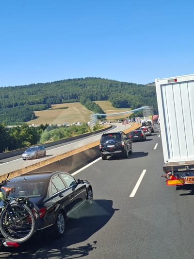 The image depicts a traffic jam on a sunny day, likely on a German autobahn near Riedenberg. The scene is dominated by vehicles stopped on the highway. In the foreground, a dark-colored car is parked, with a bicycle mounted on its rear. To its left, a silver car is also stopped. Further ahead, a black SUV is visible, followed by other cars and a white truck. The highway is bordered by a lush green forest on one side, with a grassy embankment and a blue barrier separating the road from the landscape. Overhead, the sky is clear and blue, indicating good weather. The sun casts long shadows, suggesting it is either early morning or late afternoon.
Notable details include a triangular warning sign with a red border and a symbol of a sharp curve, indicating a change in road conditions. The license plates of some vehicles are partially visible, with one reading "KRU-JB 773". The white truck has some text visible on its rear, appearing to be part of a license plate or company name, including "KR" and "IZR". The overall mood seems to be one of frustration or waiting due to the traffic congestion. The scene is dynamic, with the shadow of a bicycle on the pavement suggesting movement or a change in position of the vehicles. The presence of the bicycle on the car hints at leisure activities planned or just concluded.