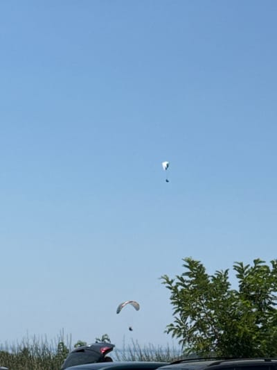 The image shows a clear blue sky with two paragliders in the air. The paragliders are at different altitudes, with one higher and further to the left, and the other lower and closer to the right side of the frame. In the foreground, the tops of several cars and green foliage are visible. The scene is likely an outdoor recreational area. The location in Boreti, Montenegro is inferred from the surrounding context of the image.