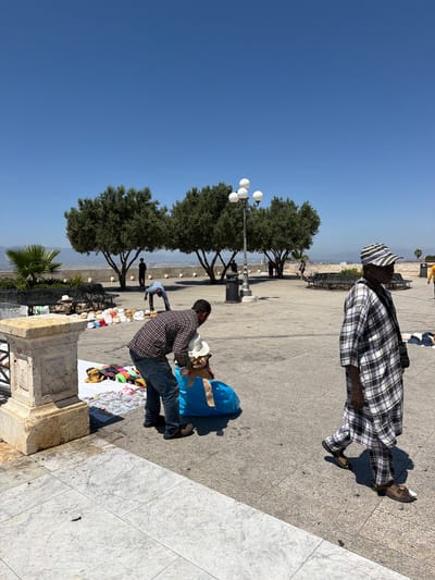 In Cagliari, Italy, on a bright sunny day, a man in a checkered outfit walks towards the right of the frame. He wears a black and white checkered shirt and matching pants, and brown sandals with white socks. In the background, another man, wearing a plaid shirt and jeans, is bent over a blue bag filled with hats and other items, apparently selling them from a makeshift stall. Hats are displayed on white sheets and on the ground. Trees and benches are visible in the background, along with a lamppost. The scene takes place on a paved square with a low stone wall on the left.