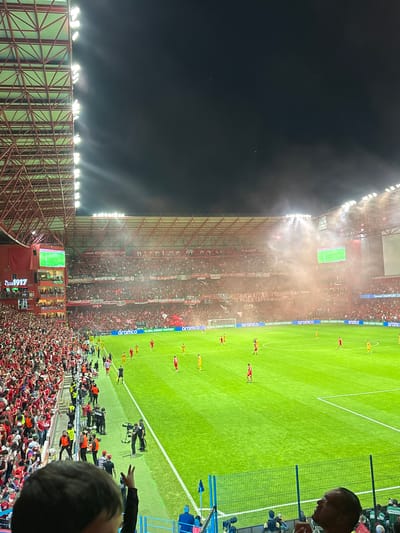 This is an image of a soccer match at the Estadio Nemesio Díez, also known as "La Bombonera," in Toluca, Mexico. The stadium is packed with fans, many wearing the team's colors, predominantly red. Some fans are setting off red smoke flares, partially obscuring the view of the field. A soccer game is in progress with players in red and yellow uniforms. Stadium lights illuminate the pitch and surrounding area. There are banners and advertisements visible. There are cameramen filming the game from the sideline. Spectators can be seen looking on. Some of them are wearing green in their hats.