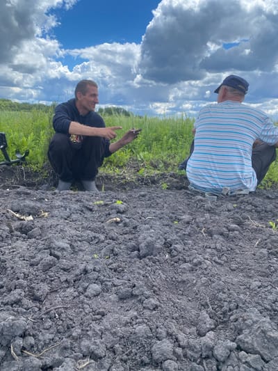 Two men are in a cultivated field near Pidluky, Ukraine. One man, wearing a striped shirt and cap, sits on the edge of the field. The other man squats in the field, holding an object, possibly a cigarette, in his hand. A metal detector is visible near the squatting man. The sky is visible with a few clouds, behind the men there is green grass. The field is dark and freshly turned.