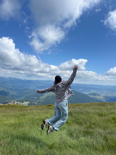 In the foreground, a person in a plaid shirt and light blue cargo pants is captured mid-jump in a grassy field. The person is wearing a dark baseball cap and athletic shoes. Their arms are outstretched, and their legs are bent. The background reveals a vast mountainous landscape with rolling green hills covered in trees. In the distance, a small village with red-roofed buildings is visible nestled among the hills. The sky above is a vibrant blue, dotted with fluffy white clouds. This scene is located in Chorna Tysa, Ukraine.