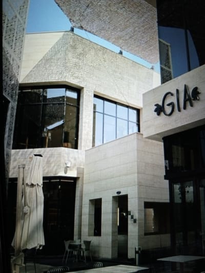 The image depicts an outdoor view of a modern building facade, featuring light-colored stone cladding and large dark-tinted windows. The building has a contemporary architectural style with geometric shapes and angled sections. Above the facade, a textured, light-colored canopy is visible, stretching across the top portion of the frame. The text "GIA" in dark, stylized lettering is prominently displayed on one section of the building, accompanied by leaf-like embellishments. Below the canopy, parts of the blue sky are visible. In the lower section of the image, outdoor seating arrangements with tables and chairs are present, suggesting a commercial establishment like a restaurant or cafe. A large, off-white umbrella or awning is partially unfurled on the left side. The scene appears to be in an urban setting.