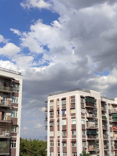 The image captures an outdoor scene featuring two apartment buildings against a backdrop of a partly cloudy sky. The buildings are tall, multi-story structures with balconies on each level. The building on the left appears to be closer to the viewer, while the building on the right is situated further back.

The apartment building on the left is characterized by its light-colored exterior, possibly white or light grey, adorned with decorative tilework in shades of brown and possibly maroon around the windows and balconies. The balconies themselves are enclosed with metal railings and some are fitted with awnings or screens. There are visible window panes, some of which appear to be open, and laundry hanging on clotheslines on some of the balconies.

The apartment building on the right shares a similar architectural style, with light-colored facades and balconies. It also displays decorative tile accents in complementary colors. Visible on its balconies are awnings and railings, and some windows are apparent.

Between the two buildings, a line, possibly a clothesline or communication wire, is stretched across the sky, connecting the structures. Below the buildings, a dense line of green trees is visible, suggesting the presence of a park, forest, or a residential area with significant greenery.

The sky is a mix of blue and grey clouds. Patches of bright blue sky are interspersed with large, fluffy white clouds and darker, more ominous grey clouds, suggesting dynamic weather conditions.

No people are clearly discernible in the image, nor are there any explicit actions or interactions being captured. The scene is a static view of urban architecture and the natural sky. The context provided about Minglola mahalla, Uzbekistan, situates this image within a specific geographical and cultural setting, implying the architectural style might be typical of residential buildings in that region.
