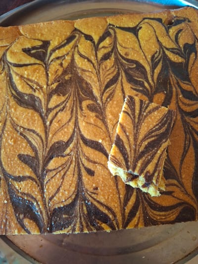 The image shows a rectangular marble cake with a swirled pattern of dark and light batter. A slice of the cake, with a bite taken out of it, is placed on top of the larger cake. The cake is on a metallic platter. The scene is indoors, likely a kitchen. The location in Ko`kdala, Uzbekistan is not discernible from the image.