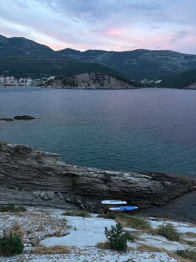 This media file depicts a coastal scene at dusk in Nerin, Montenegro. The image is oriented vertically, with the foreground dominated by a rocky cliff face. Two paddleboards are resting on a narrow ledge on the cliff. One paddleboard is predominantly blue with white accents and a red fin, while the other is white with a patterned design.
Beyond the cliff, a calm, dark blue body of water stretches out. In the distance, a town or village with numerous buildings is nestled along the base of a steep, forested mountain range. The mountains rise dramatically from the coastline, their slopes covered in dense vegetation.
The sky above the mountains is transitioning from daylight to evening, with soft hues of pink and purple visible, suggesting a sunset or twilight. Wispy clouds are scattered across the sky. The overall atmosphere is serene and tranquil. No people are visible in the scene. The time of day is clearly evening, and the weather appears clear and calm. The location is identifiable by the mountainous coastline and the architecture of the distant settlement, consistent with coastal towns in Montenegro.