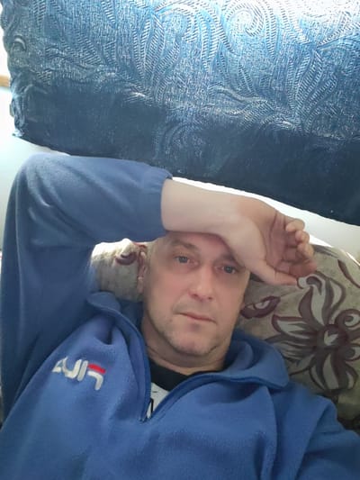 This indoor portrait features a middle-aged man with light hair, blue eyes, and light stubble, lying down in a relaxed or contemplative pose. He is wearing a blue fleece jacket with a visible zipper, sporting a white and red "FILA" logo on the left chest, which appears stylized or slightly reversed. His head rests on a patterned cushion adorned with brown and beige floral or leaf-like motifs. His right arm is bent, with his hand placed on his forehead, suggesting either relaxation, weariness, or thought. In the background, above him, is a dark blue textured fabric, possibly another cushion or furniture upholstery, displaying an intricate, woven or embroidered pattern. The lighting is soft, indicating an indoor setting. The image is set in Rybinsk, Russia.