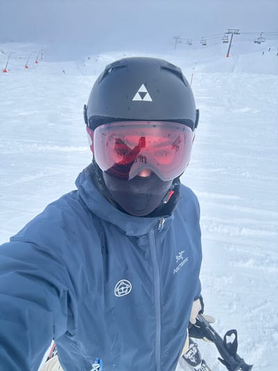 An individual is depicted in a close-up, chest-up selfie on a snow-covered ski slope in Bad Hofgastein, Austria. The person wears a black ski helmet with a white triple-triangle logo on the forehead, and large red-tinted ski goggles. The goggles reflect a snowy mountain landscape, featuring indistinct figures, likely other skiers, and ski lift structures in the distance. A black balaclava covers the lower face, visible beneath the goggles. The individual wears a blue long-sleeved ski jacket, featuring an "ARC'TERYX" logo on the right chest and a circular white emblem on the left arm. A light-colored gloved hand grasps a black ski pole visible in the lower right of the frame, with a portion of white skis present in the lower left. The background consists of an expansive, groomed ski run ascending into an overcast, hazy sky. Multiple chairlifts are visible traversing the mid-ground, and orange marker poles line the left side of the slope, defining a boundary. The overall environment is a ski resort under cloudy weather conditions.