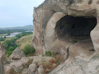 The image shows a view from within a cave-like opening carved into a large rock formation. The opening is arched and the interior is dark and textured. Outside the opening, in the foreground, are large, weathered rocks with sparse vegetation. In the mid-ground, a river winds through a valley with green trees and rolling hills. In the background, distant hills meet a hazy sky. The scene is located in Uplistsikhe, Georgia, an ancient rock-hewn town.