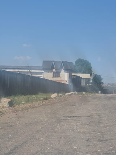 The image depicts an outdoor scene with a dusty, unpaved road in the foreground. Beyond the road, a wooden fence runs horizontally across the frame, with grass and a few rocks situated between the fence and the road. Behind the fence, there is a white brick building with a grey roof, featuring two prominent gables. To the right of this building, a lighter-colored, single-story structure with a light grey roof is visible, followed by dense green foliage. Smoke is emanating from behind the fence and the buildings, obscuring parts of the scene and drifting upwards and towards the right. The sky above is clear and blue, with a few wispy clouds. The overall impression is of a rural or semi-industrial area, possibly a residential backyard or industrial lot, on a sunny day with evidence of smoke. It is not possible to determine the specific location within Pidhaitsi, Ukraine from the image alone, as no identifying landmarks or signs are visible.