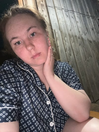A young Caucasian woman with fair skin, freckles, and light brown hair is pictured from the chest up, with her face turned towards the camera. She is wearing dark blue pajamas with a white polka dot pattern and white piping. Her right hand is gently touching her cheek and jawline, and she is looking directly into the camera with a neutral expression. The background consists of a weathered wooden wall, suggesting an outdoor or rustic indoor setting, possibly in a rural area like Аряж, Russia. The lighting is dim, creating shadows and highlighting the textures of the wood and skin.