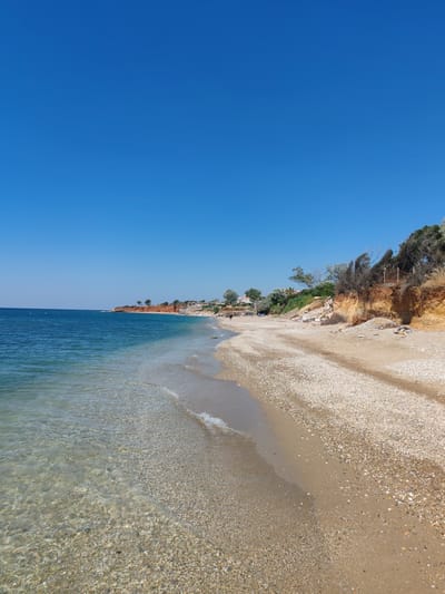 The image depicts a view of a beach in Alexandroupolis, Greece, characterized by its clear blue sky, calm sea, and a sandy and pebbly shoreline. The water is translucent, allowing visibility of the seabed. The coastline features a reddish-brown cliff face on the right side, dotted with sparse vegetation. Several buildings, possibly houses or tourist accommodations, are visible on the cliff's edge and further inland. A few individuals are present on the beach, some walking near the water and others relaxing further up the shore. Beach umbrellas and a few trees add to the scene. The overall impression is a peaceful coastal environment under a bright, sunny sky.
