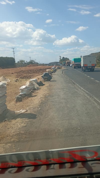 The image captures a wide outdoor scene on a bright, sunny day, likely taken from inside a moving vehicle.

On the left side of the frame, there is an unpaved, dusty ground with sparse dry grass. Along the immediate edge of this dirt area, running parallel to the road, are numerous bags, likely containing cement or sand, stacked in piles. Most bags are white or off-white, and some are covered by a dark, possibly black, tarp or plastic sheeting. Further back, the ground is disturbed, showing reddish-brown earth, indicating recent excavation or construction work. In the mid-distance on the left, a line of green bushes or small trees is visible, with utility poles and power lines extending across the landscape. Distant hills or elevated terrain can be seen on the horizon.

The center and right side of the image are dominated by a two-lane asphalt road. The road surface appears well-maintained, with a dashed white line separating the two lanes. A significant amount of traffic, primarily large trucks and some smaller vehicles, is visible on the right lane, stretching into the distance and appearing to be in a queue or moving slowly. The trucks vary in appearance; some have open beds covered with green tarps, while others are enclosed box trailers, one of which is light-colored with visible rear markings. Beyond the road on the right, more hilly and vegetated terrain is visible in the background.

The sky is a clear, bright blue with scattered white, puffy cumulus clouds. The lighting suggests it is daytime, probably mid-morning or afternoon, given the strong shadows.

At the very bottom of the image, the interior of the vehicle from which the photo was taken is partially visible. There's a reflective surface, possibly a dashboard or a window reflection, displaying reversed and slightly distorted red lettering. The visible text appears to spell out "YETU", reflected upside down and backwards, in a bold, stylized font.