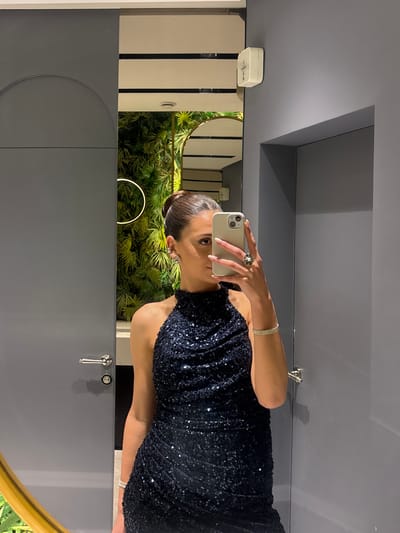 A woman is standing in a modern bathroom, taking a selfie in the mirror with her iPhone. She is wearing a sleeveless, dark blue dress with sequins, a ring on her left hand, and a bracelet on her right wrist. The bathroom has gray walls, and in the mirror's reflection, there is a green plant. The location is most likely a private residence in the city of Masis, Armenia.