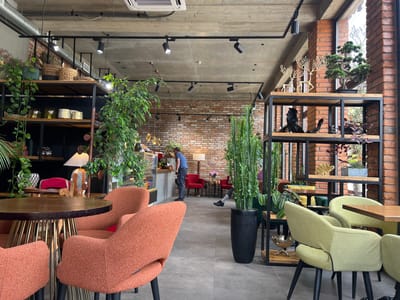 The image depicts the interior of a contemporary cafe in Adzhiryuara, Abkhazia. The space is characterized by exposed brick walls, a concrete ceiling with visible ductwork and track lighting, and industrial-style shelving units. Numerous plants, including large potted ferns and a bonsai tree, are strategically placed throughout the cafe, contributing to a biophilic design.
Seating areas feature a mix of furniture: several plush, coral-colored armchairs are arranged around a circular wooden table with a metallic base in the foreground. In the background, there are two red upholstered chairs. Further into the cafe, lime-green armchairs are positioned around rectangular wooden tables.
A man in a blue t-shirt and dark pants is visible near a counter, appearing to be an employee, possibly attending to a customer or preparing orders. Another person's legs and feet are visible seated on one of the green chairs. The counter area shows display cases with food items and decorative elements.
Decorative objects include golden reindeer figures, ceramic vases, and sculpted wood pieces placed on shelves. The overall atmosphere suggests a modern, stylish, and comfortable cafe environment.
