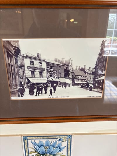A black and white historical photograph depicts "The Cross, Oswestry, United Kingdom," a bustling street scene. The image features a wide street lined with multi-story commercial buildings. On the left, a prominent building houses "FRANCIS & BECKITT WHOLESALE & RETAIL GROCERS," displaying signs for "TEA & COFFEE EXPERTS" and "HIGH-CLASS PROVISIONS." Adjacent structures include "ANDERSON" and "PEOPLES' FOOT MART," with a sign indicating "FRUITS IN SEASON." Further down the street, buildings exhibit traditional half-timbered architecture. The street and sidewalks are populated with numerous pedestrians, including men, women, and children, all dressed in late 19th or early 20th-century clothing. Men are typically seen in suits and various hats (bowlers, flat caps), while women wear long dresses and elaborate hats. Individuals are depicted walking, congregating in small groups, or standing near shopfronts, creating a lively urban tableau. The photograph is labeled "58690" and "VALENTINES SERIES" at the bottom.