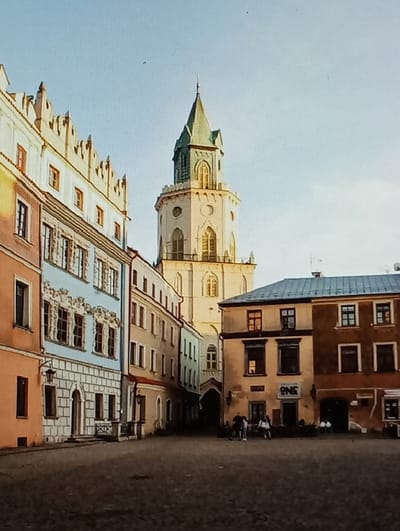 The image displays a daytime view of the Rynek (Market Square) in Legnica, Poland. Dominating the background-center is the Old Town Hall tower (Stary Ratusz), a multi-tiered, light-colored structure with gothic-style arched windows, topped by a pointed green spire.
To the left, a row of multi-story historic tenement buildings lines a street or passage. The building closest on the left features an orange-brown base, a light blue mid-section with ornate window surrounds, and a white upper level with a decorative stepped gable. A black wall-mounted lantern is visible on its lower facade. Further down this row, buildings with simpler beige and light brown facades recede.
On the right side of the frame, two multi-story buildings face the square. The left building is light brown/ochre with dark windows and a sign reading "ONE" above an entrance. The adjacent building to its right is darker brown.
The foreground consists of a dark, paved square, likely cobblestone, with visible textural variations and patches of shadow extending from the right, suggesting directional sunlight. A small group of approximately three to five indistinct figures is present near the base of the buildings on the right, possibly interacting or seated. The sky is clear and light blue.