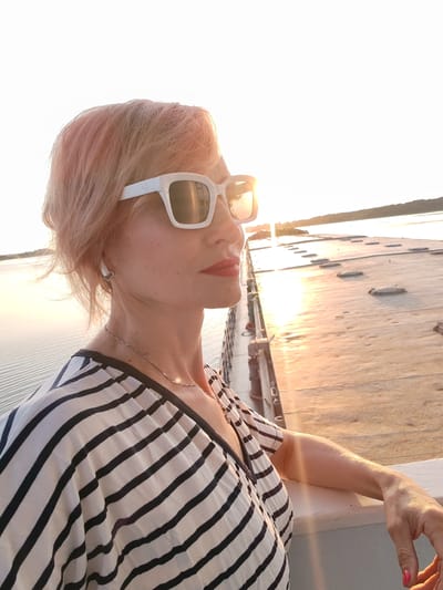 A woman with blonde hair and white sunglasses is captured in a close-up, side-profile shot as she looks out over a body of water. She is wearing a black and white striped top and a delicate necklace. Her right arm is resting on a white railing. The background features a large, flat structure, possibly a deck, illuminated by the golden light of the setting sun, which also casts a warm glow on the water and the distant treeline. The sky is bright and clear, indicating an outdoor setting, likely during the golden hour. The location is identified as Shchetinino, Russia.
