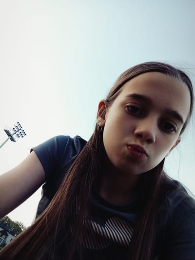 The image is a selfie taken from a low angle, with the subject being a young person. The person has long, dark brown hair that falls to the side of their face. They are wearing a dark t-shirt with a striped pattern on the chest. The background is a bright, overcast sky, with a lamppost and some buildings visible in the upper left corner. The person's face is turned towards the camera, with their eyes looking directly at the viewer. Their lips are slightly parted, giving them a pensive expression. The lighting suggests it is daytime, possibly late afternoon, given the soft, diffused light. The environment appears to be outdoors, likely in a park or open area, considering the presence of a lamppost which resembles a sports stadium light. There is no visible text in the image. The scene is static, with the subject posing for the selfie. The emotional tone of the image is neutral to slightly introspective. The location context provided is Radzymin, Poland.