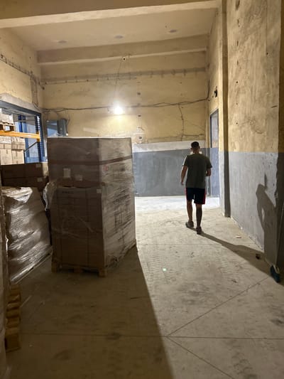 This media file captures an indoor scene within what appears to be a warehouse or unfinished building. The setting is characterized by raw concrete floors and walls, with exposed electrical conduits running along the walls. In the foreground, several large cardboard boxes, some wrapped in clear plastic, are stacked on wooden pallets. One stack of boxes prominently displays the text "MARIO".
A person, seen from the back, is standing in the middle ground. They are wearing a dark green t-shirt and black shorts with red trim, and casual shoes. The person's posture suggests they are looking around the space. The lighting is somewhat dim, with a bright overhead light illuminating a section of the floor and casting a long shadow.
The overall impression is one of an industrial or storage area, possibly under construction or in the process of being organized. The time of day is indeterminate due to the indoor setting. There are no visible external weather cues. The location is identified as Djerman, Bulgaria. No specific emotions are conveyed, but the scene suggests a practical, functional environment.
