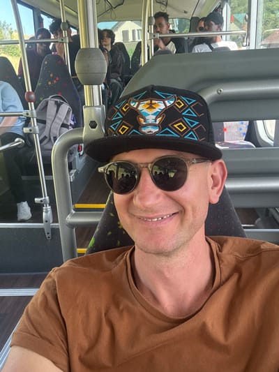 A close-up, selfie-style photo is taken from inside a bus in Saicourt, Switzerland, by a man with fair skin and a light beard wearing sunglasses and a brown t-shirt. He is also wearing a black baseball cap with a bull design on it. The man is smiling and looking at the camera. In the background, other passengers are visible on the bus, as well as the interior of the bus with seats and handrails. The windows of the bus show a blurred view of the outside, with trees and buildings visible. The lighting suggests it is daytime.