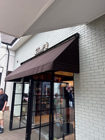 The image depicts the exterior of a shop, likely a chocolatier, identified by the name "Maldon" on the awning. The building has a white brick facade and a brown awning. A glass storefront displays various confectioneries, including what appears to be chocolate truffles and other sweets. The reflection in the glass shows the photographer and a man wearing a black t-shirt and khaki shorts standing outside the shop. The doors are open, revealing the interior. The location appears to be on a brick-paved street. The city is Braintree, United Kingdom.