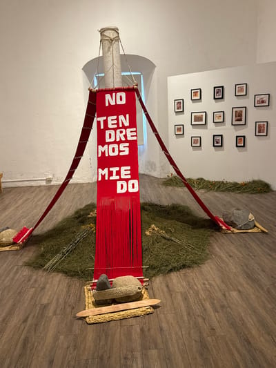 An art installation dominates the room. A red banner with white text "NO TEN DRE MOS MIEDO" is suspended by red supports from a white column to platforms on the wooden floor. Green foliage surrounds the platforms, each anchored by a large rock. A collection of framed photos hangs on the white wall in the background.