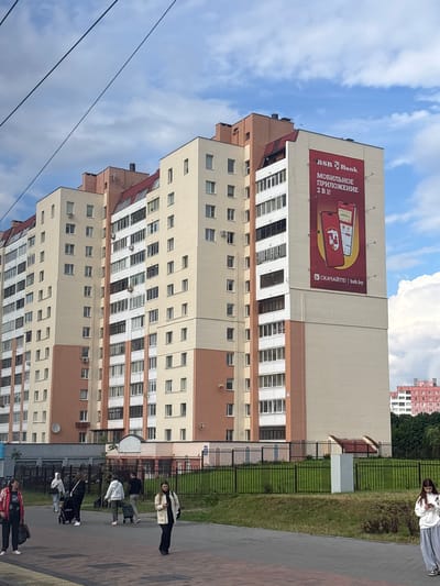 A large apartment building with cream and red facades dominates the frame. A prominent red billboard advertising "BSB Bank Mobile Application" is attached to the upper section of the building. The billboard features images of two smartphones and text in Cyrillic. In the foreground, a paved walkway runs along a grassy area bordered by a dark metal fence. Several people are walking on the walkway. To the left, a woman in a red jacket and black pants carries a large black bag. Behind her, a person pushing a stroller walks with another person. Further to the right, another individual pushes a rolling cart. On the far right, a young woman in a white hoodie and grey pants looks at her phone. The sky above is a mix of blue and scattered white clouds. In the background, other buildings and trees are visible. The overall scene appears to be a residential area in the vicinity of Сеніцкі сельскі Савет, Belarus.