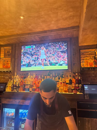 The image depicts the interior of a bar or restaurant, with a large screen displaying a tennis match, presumably Wimbledon, given the context of the player's attire and the court. A bartender, wearing an apron over a black t-shirt, is visible in the foreground, looking down at something on the bar. The bar itself is stocked with a wide array of liquor bottles, and there is a refrigerated display unit containing canned beverages and bottles of drinks. The overall atmosphere suggests a lively establishment. The question of the location in Peterborough, United Kingdom, cannot be answered from the visual information provided in the image.