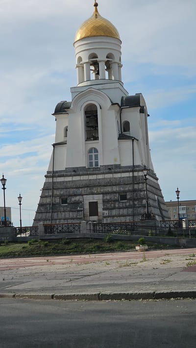 The image shows a bell tower in Kamensk-Uralsky, Russia. The bell tower is white with a golden dome and has a square base. There are bells visible in the upper section of the tower. The tower is surrounded by a fence and some vegetation. In the background, there are buildings and a cloudy sky. The scene appears to be a public space with a paved area in front of the tower.