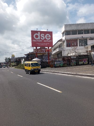 The image depicts a daytime street scene, identified as being in the city of Gubbisamamidi, India, under an overcast sky. A two-lane asphalt road with solid and dashed white markings extends into the distance. A yellow van, traveling away from the viewer, occupies the right lane in the foreground. Multiple vehicles, including motorcycles, are visible further down the road on the left.
On the right side of the road, a prominent red billboard displays "dse FACTORY OUTLET" in white lettering, with the address "Jl. Raya Cimacan 39 Puncak" and telephone number "(0263) 511787" below it. A smaller red sign beneath the main billboard advertises "BIG SALE 70%." Adjacent to the billboard stands a multi-story commercial building with a light-colored, weathered exterior. Its upper floor windows showcase clothing on mannequins, and a satellite dish is attached to the facade. The ground level features several vendor stalls, one displaying green coconuts. Multiple individuals are present on the sidewalk in front of these stalls. Additional commercial structures, utility poles, and signage line the street in the background.