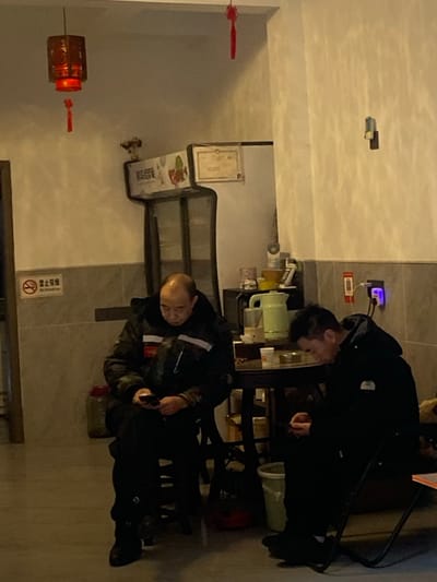 Two men in dark clothing are present in a dimly lit interior. One stands near a table with a green kettle and other items, while the other is seated with what appears to be a tool in his hand. Decorative lanterns hang on the left. Signs, possibly in Chinese, are visible on the wall. The room's walls are a light color.
