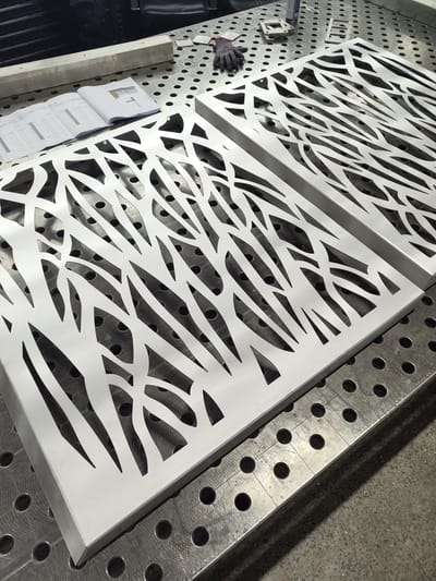 Two laser-cut metal panels with an organic, branch-like pattern are positioned on a metal grid table. A technical drawing, a grey glove, and other metal pieces rest nearby. The environment appears to be a metal fabrication or workshop space. I have no information about the location in the city of Manternach, Luxembourg.