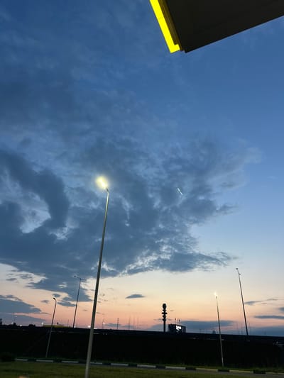 A wide-angle, low-angle shot captures an overcast sky at dusk above a suburban landscape in Budovo, Russia. The sky is a gradient of deep blues and grays, with patches of lighter clouds illuminated by the fading sunlight near the horizon. A bright yellow stripe runs along the top edge of the frame, hinting at a structure above.

In the foreground, a tall, slender streetlamp with a glowing white bulb stands prominently on the left side of the image. To the right, a darker, more distant streetlamp rises diagonally.

Below the sky, a dark, dense line of trees or bushes forms a silhouette against the colorful horizon. Scattered lights from distant buildings and streetlights dot the landscape. A large, dark rectangular structure with illuminated signs is visible in the middle distance.

The ground in the immediate foreground is a grassy area with a paved path or road. The overall impression is one of a quiet, transitional moment at the end of the day in a somewhat industrial or commercial area. No people are visible in the frame.