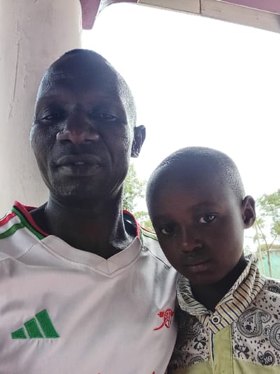 A close-up shot features two dark-skinned individuals, an adult male and a child. The adult wears a white t-shirt with green and red accents and a small red logo. The child is dressed in a patterned shirt with button details. The camera angle looks up at them, with bright light washing out the background. Their faces are clearly visible, both looking toward the camera. The adult is positioned above the child.