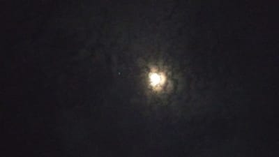 The image depicts a night sky scene in Cotbadatunong, Indonesia, dominated by a bright, partially obscured moon. The moon is the central subject, appearing as a luminous orb surrounded by wispy clouds that create a halo effect. To the left of the moon, a smaller, distinct green object is visible, possibly a distant light or celestial body. The overall atmosphere is dark, indicative of nighttime, with the clouds adding a sense of depth and texture to the sky. No people or specific structures are visible, suggesting an outdoor, possibly rural, setting. The image captures a quiet and serene moment in the night, with the celestial display taking center stage. The time of day is clearly night, and the presence of clouds suggests it is not a perfectly clear night. The location is identified as Cotbadatunong, Indonesia. There is no visible text in the image.
