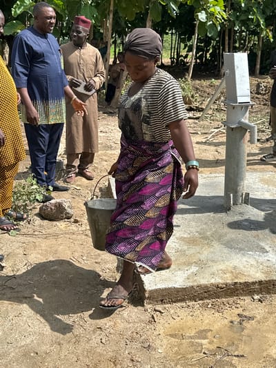 In Afogo, Nigeria, a group of people gather near a water pump. A woman, wearing a striped shirt and a colorful patterned skirt, is in the foreground, carrying a metal bucket. She stands on a concrete structure that is part of the water pump system. To her left, a man in a blue garment and a man in a brown robe are conversing. Behind them, the environment is rural, with trees and vegetation indicating a natural setting. The ground is dusty and dry, with some puddles of water visible. The water pump itself is a simple, manual model, suggesting it is a communal resource. The overall scene depicts a moment of everyday life and resource gathering in a rural Nigerian community.