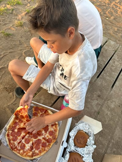 In Kostovaty, Russia, a boy with short brown hair sits on a wooden picnic table, reaching for a slice of pepperoni pizza from an open pizza box. He wears a light-colored t-shirt with a graphic print and light-colored shorts. Next to the pizza box is another open box containing a hamburger wrapped in aluminum foil. The ground around the table is sandy, with patches of dry grass. The setting appears to be outdoors, possibly a park or recreational area.