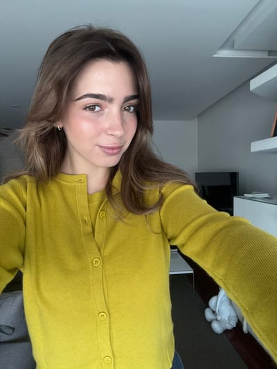 A light-skinned female subject is depicted in a close-up selfie within an indoor modern residential setting in Nogueira e Silva Escura, Portugal. She possesses light brown, shoulder-length hair styled with a middle part, brown eyes, and pronounced eyebrows. Her facial expression displays a subtle smile. She wears small hoop earrings. Her attire consists of a mustard yellow knitted long-sleeved cardigan, unbuttoned, worn over a matching short-sleeved top. The cardigan features four visible circular yellow buttons down the front. The background comprises light grey walls and a similarly colored ceiling. To the upper right, a white floating shelf is mounted, beneath which a black flat-screen television rests on a low white cabinet. A grey fabric cushion is partially visible on the left side of the frame. On the dark floor, in the lower right, a small white decorative figurine resembling a bunny is present. The subject's right arm is extended, holding the camera to capture the image.